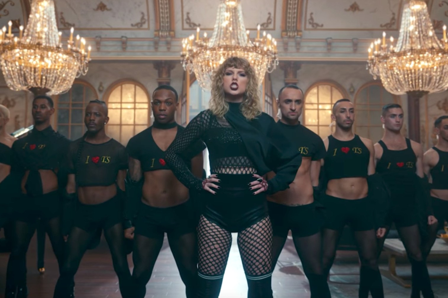 Taylor Swift in front of a row of dancers, all wearing a black version of the I Heart TS T-shirt.