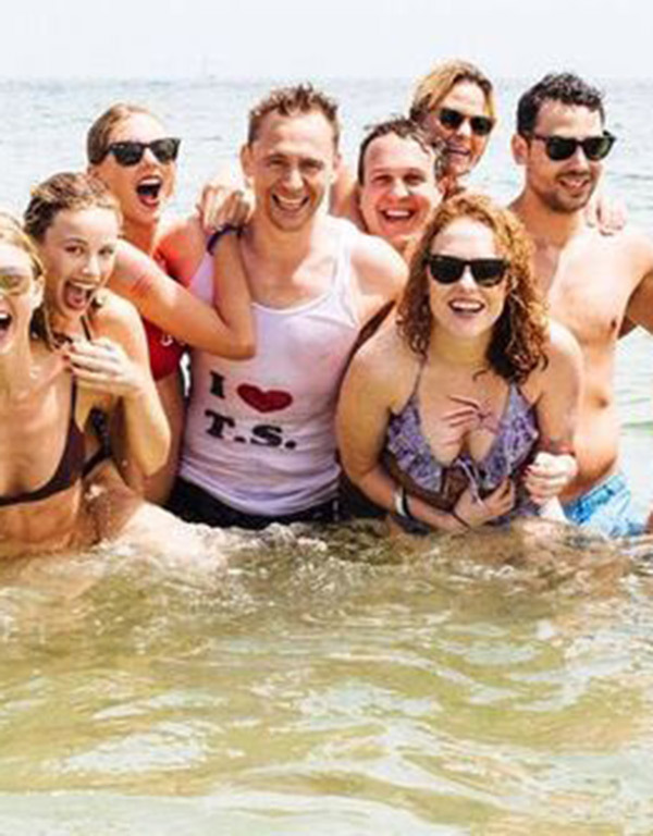 Taylor Swift and Tom Hiddleston surrounded by a group of friends at the beach; Tom is wearing a T-shirt with the logo "I Heart TS."