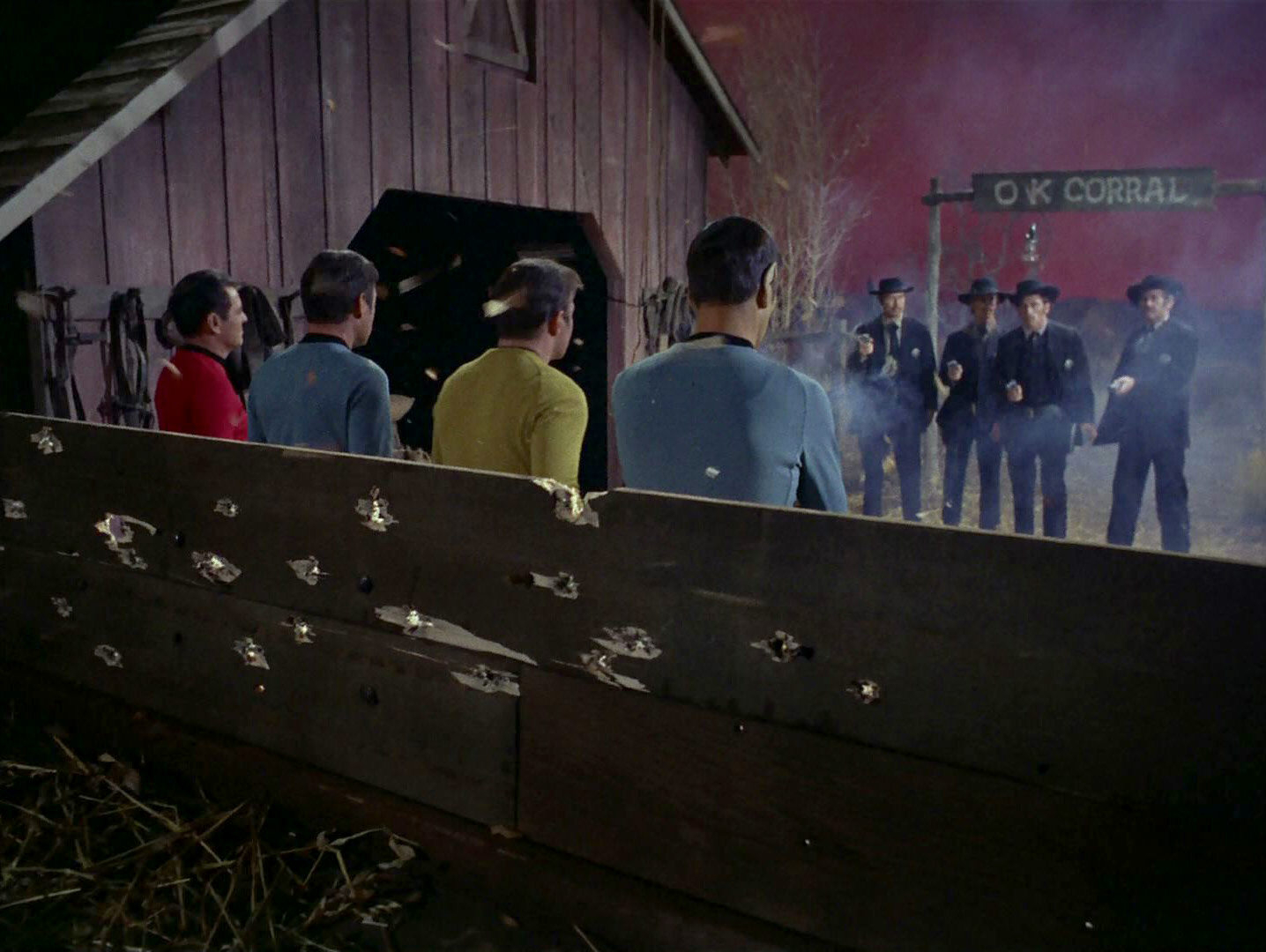 A still from Star Trek episode 'Spectre of the Gun', showing four Star Trek crew members from behind (Spock is closest to the camera, with Kirk to his left), backs against some wooden boards which are riddled with bullet holes. Across from them, four cowboys stand with guns raised, firing at them. A sign above their heads reads 'OK CORRAL'.