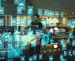 A man in a laboratory stands behind a semi-transparent wall of glowing holographic images and uses his hands to manipulate them.