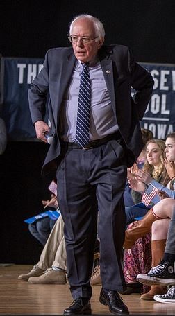 Bernie Sanders wearing a wrinkled suit and tie in front of clapping high school students.