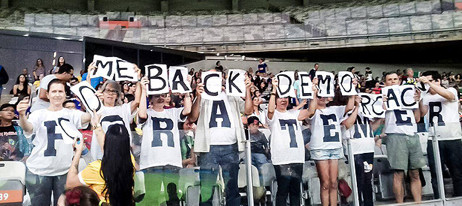 The same group of sports fans as in the previous image, this time standing up and holding smaller signs that combined read Come Back Democracy.