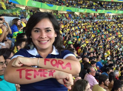 A woman stands against a background of a large audience at a sporting event. Her arms are crossed in front of her, with Fora Temer written in red on her forearms.