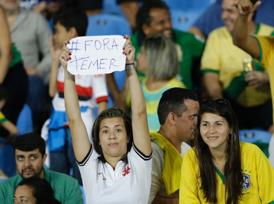 A woman standing up in the audience to a sporting event, holding up a piece of paper with #Fora Temer written on it.