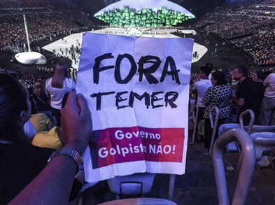 A fan in the upper rows of a stadium holds a handwritten poster up against the view of the field, with the words Fora Temer; Governo Golpisto Nao!