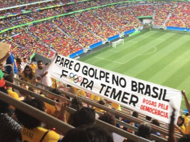 The view of a sports field from the upper levels of the stadium, showing a group of fans holding a banner reading Pare O Golpe No Brasil. Fora Temer Rosas Pela Democracia. The letter o in Fora is replaced by the logo of the Olympic rings.