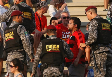 Security guards wearing camouflage and vests identifying them as Forca nacional escort a man in a red Flamengo T-shirt from the bleachers of a sporting stadium.