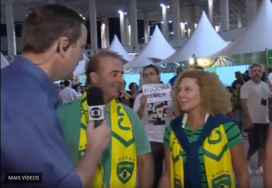 Two sports fans are interviewed by a reporter while in the background another fan holds up a poster reading Fora Temer, with the Olympic rings logo in place of the letter o.