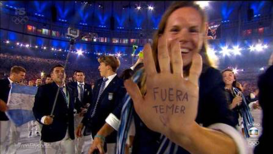 An athlete parading on the field holds her hand palm out to the camera; written on her palm in ink is Fuera Temer.