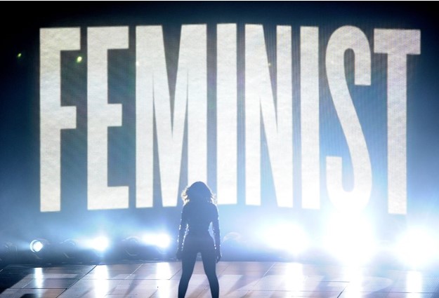 Color image of Beyoncé, in shadow, standing on stage in front of the giant word 'FEMINIST.'