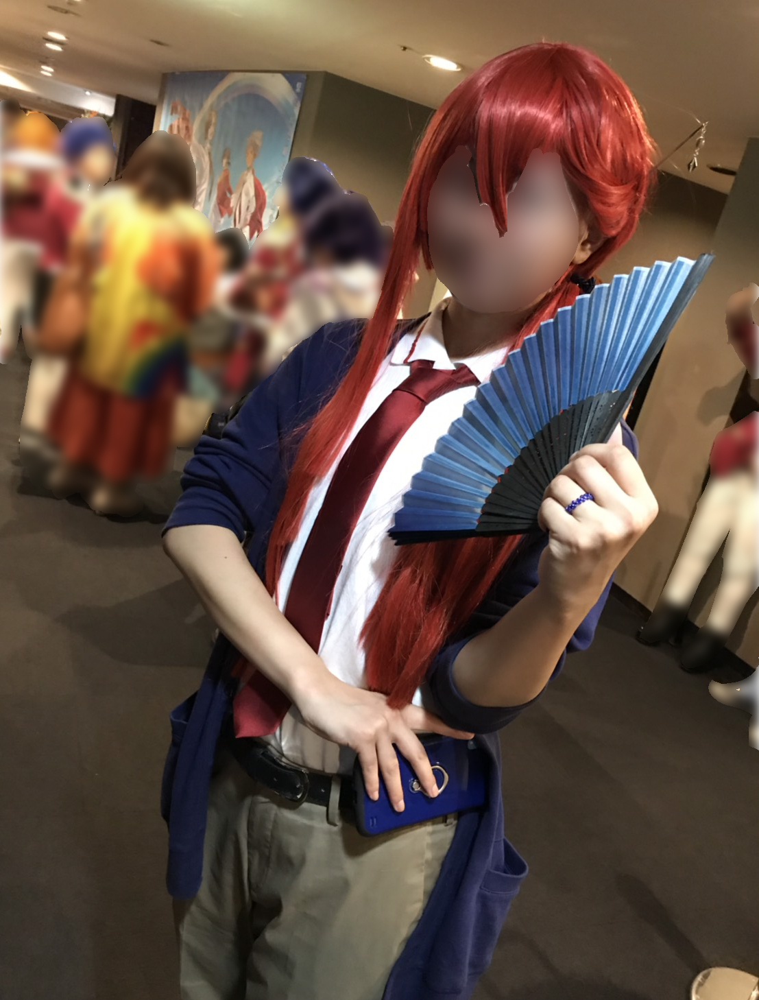 A fan whose face is blurred dressed in a red wig, white shirt, red tie, blue sweater, and tan pants, holding a fan.