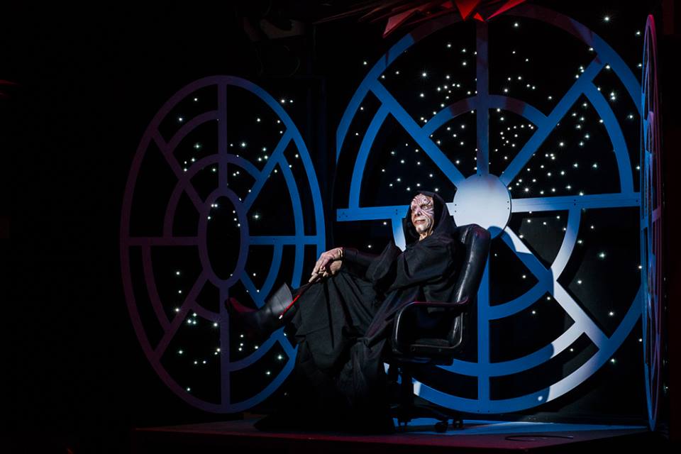 Production photo of Palpatine posing, legs-crossed in an office chair, in front of the Death Star viewport.