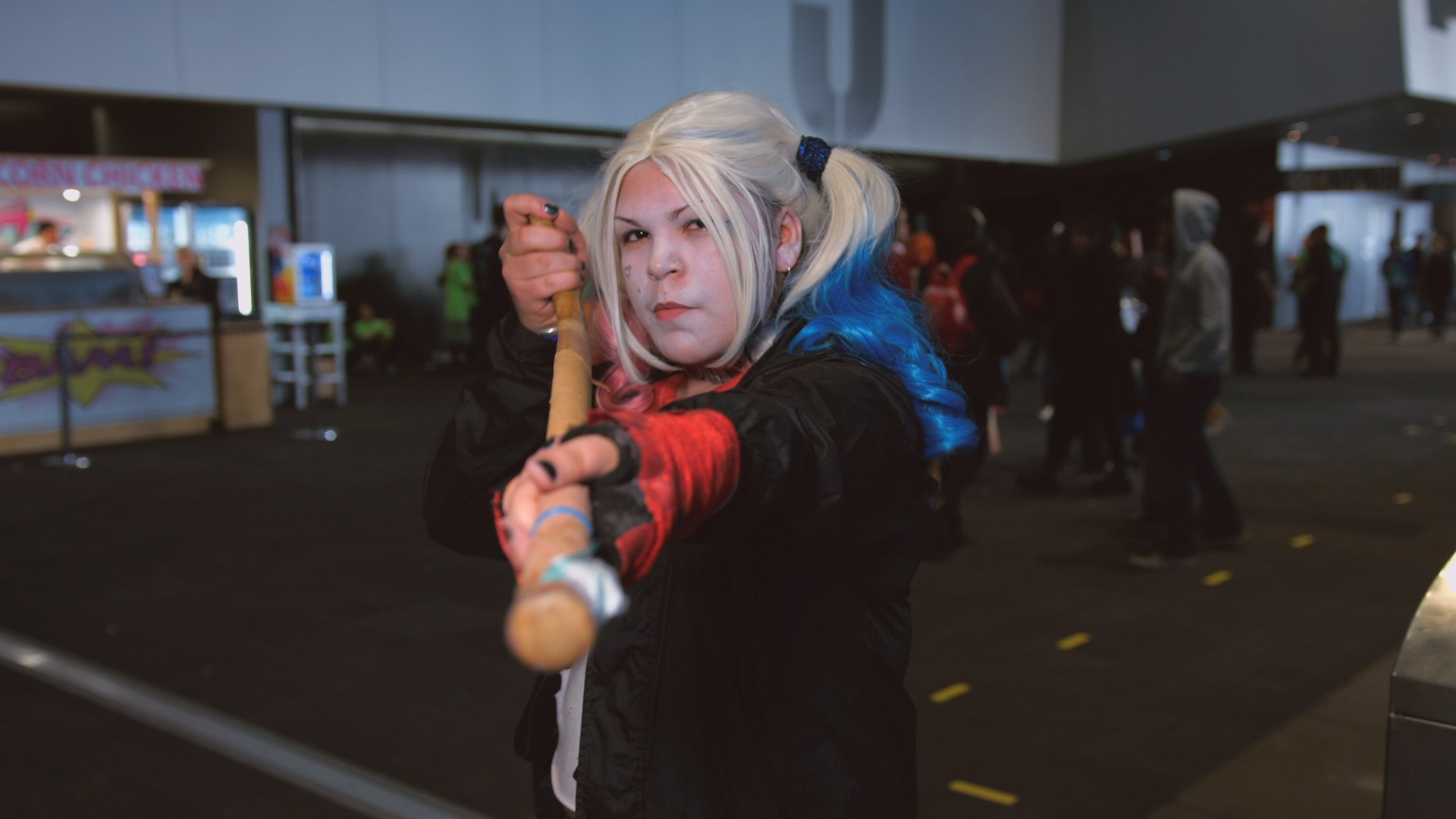 Harley Quinn cosplayer aiming a field hockey stick like a long gun.