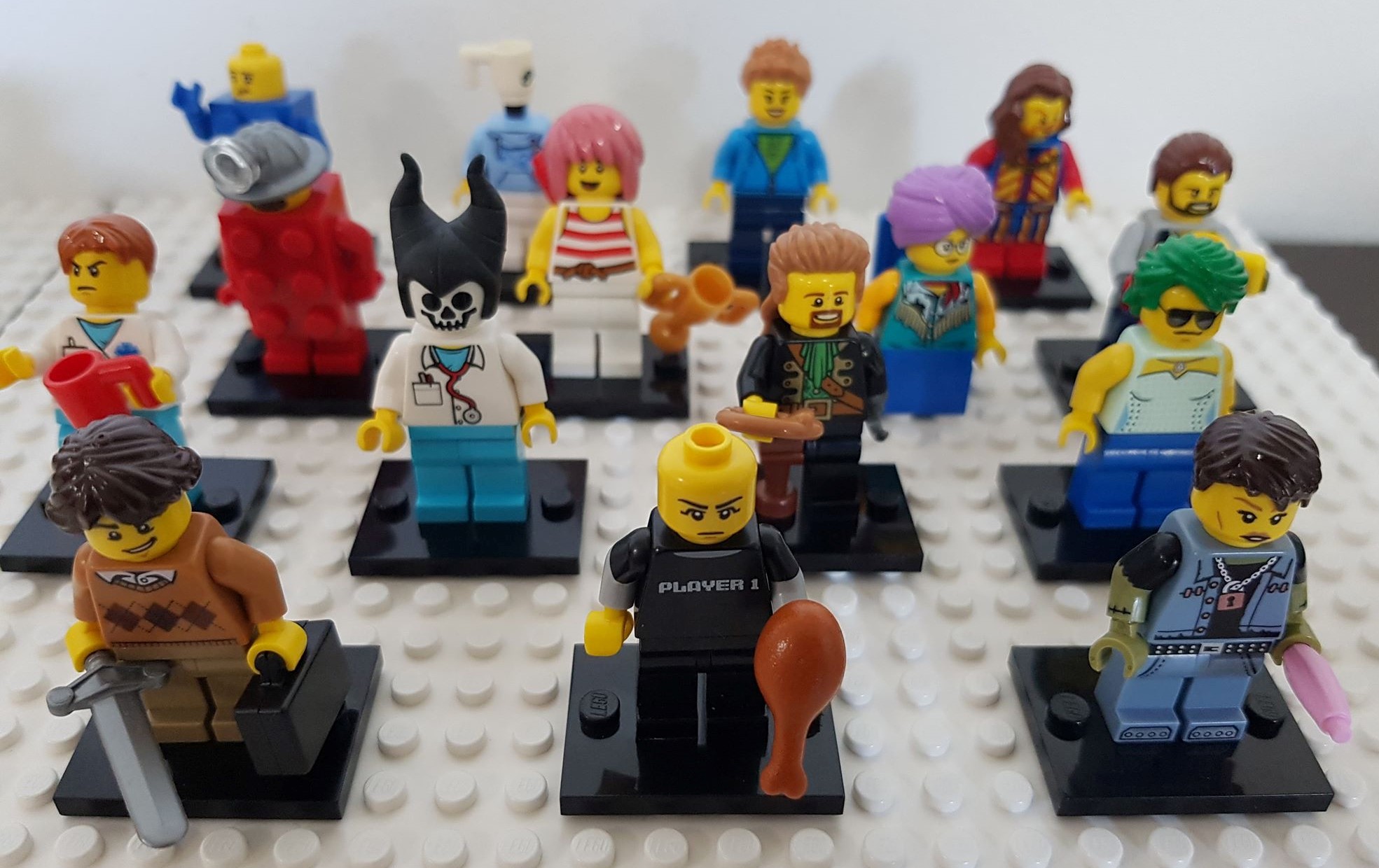 Photograph of 15 minifigures arranged in rows on a Lego baseplate. The figures have a variety of different hairstyles, heads and torsos including one which simply has a cup for a head, one which resembles a ghoul with a lab coat and stethoscope, and a bald figure wearing a shirt that reads Player 1. A few are holding items such as a sword, a trophy and a pink rolling pin.
