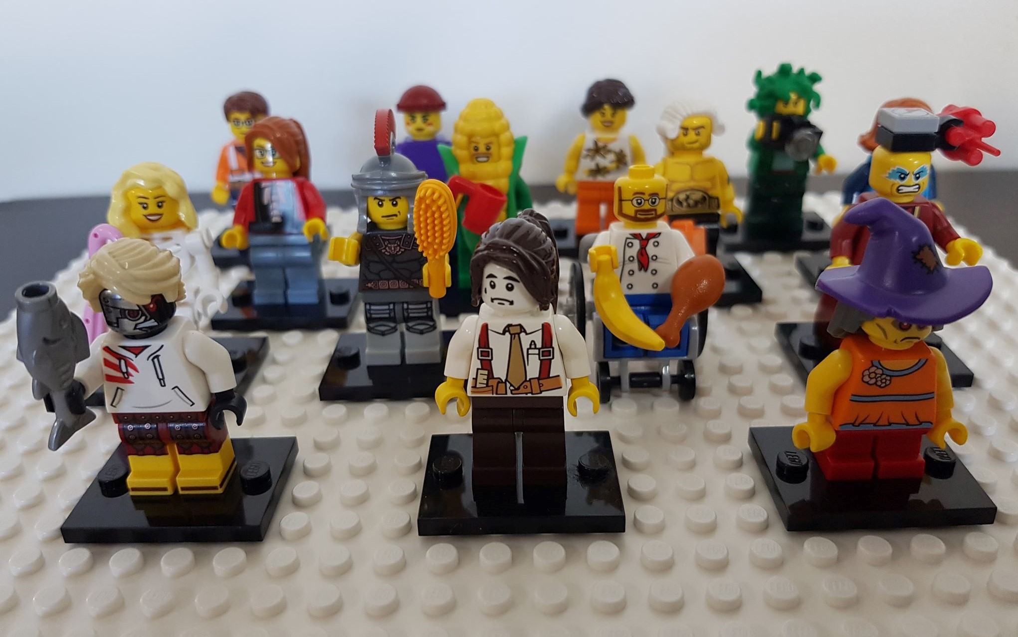 Photograph of 15 minifigures arranged in rows on a Lego baseplate. The figures are composed of a mishmash of head and body parts such as a Roman centurion, a corn cob, a Medusa and a judge's wig on a wrestler's body, and many are also holding accessories such as a banana, chicken leg or hairbrush.
