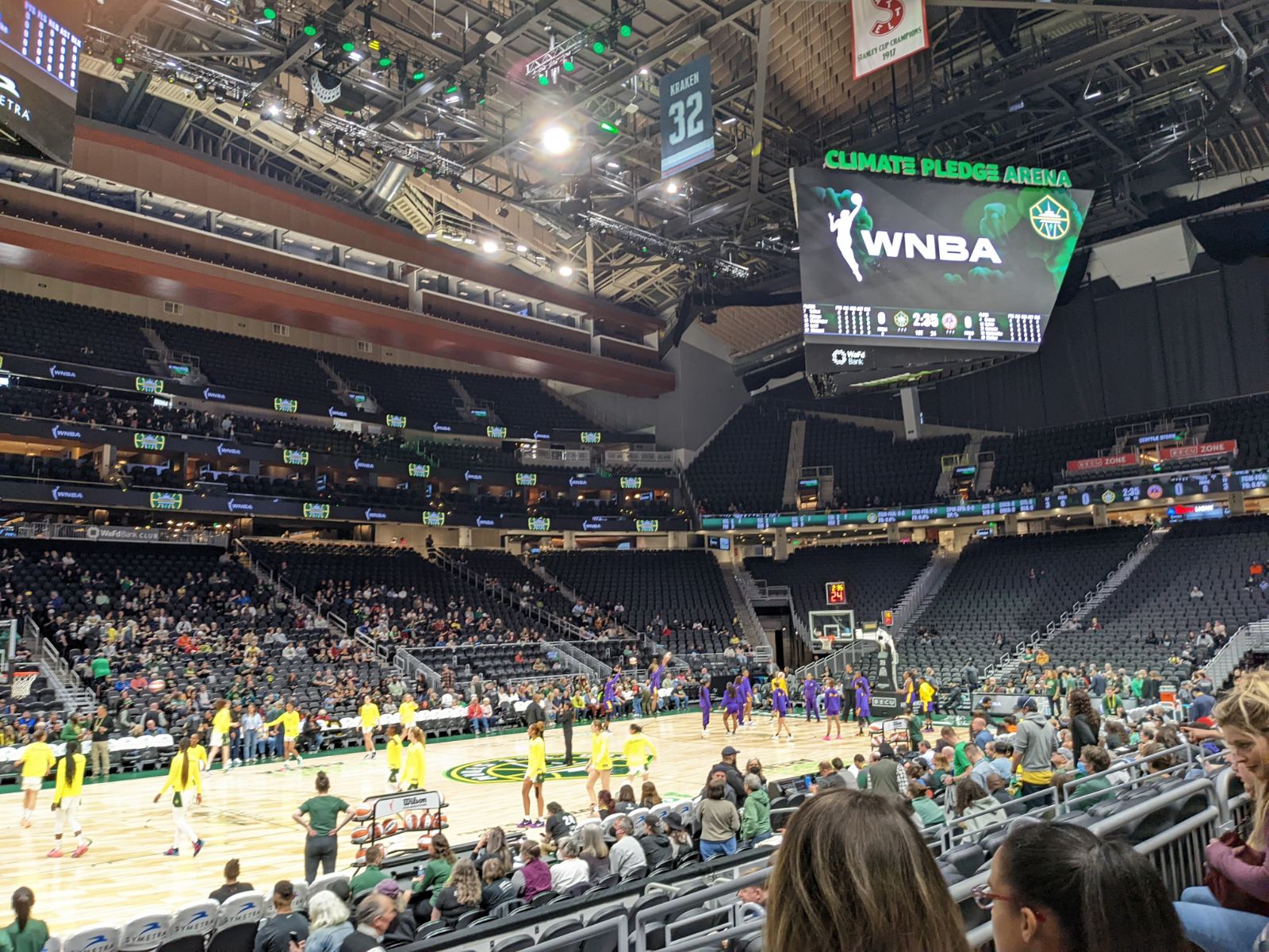 Basketball players in yellow shirts under a Jumbotron screen with the stadium name.