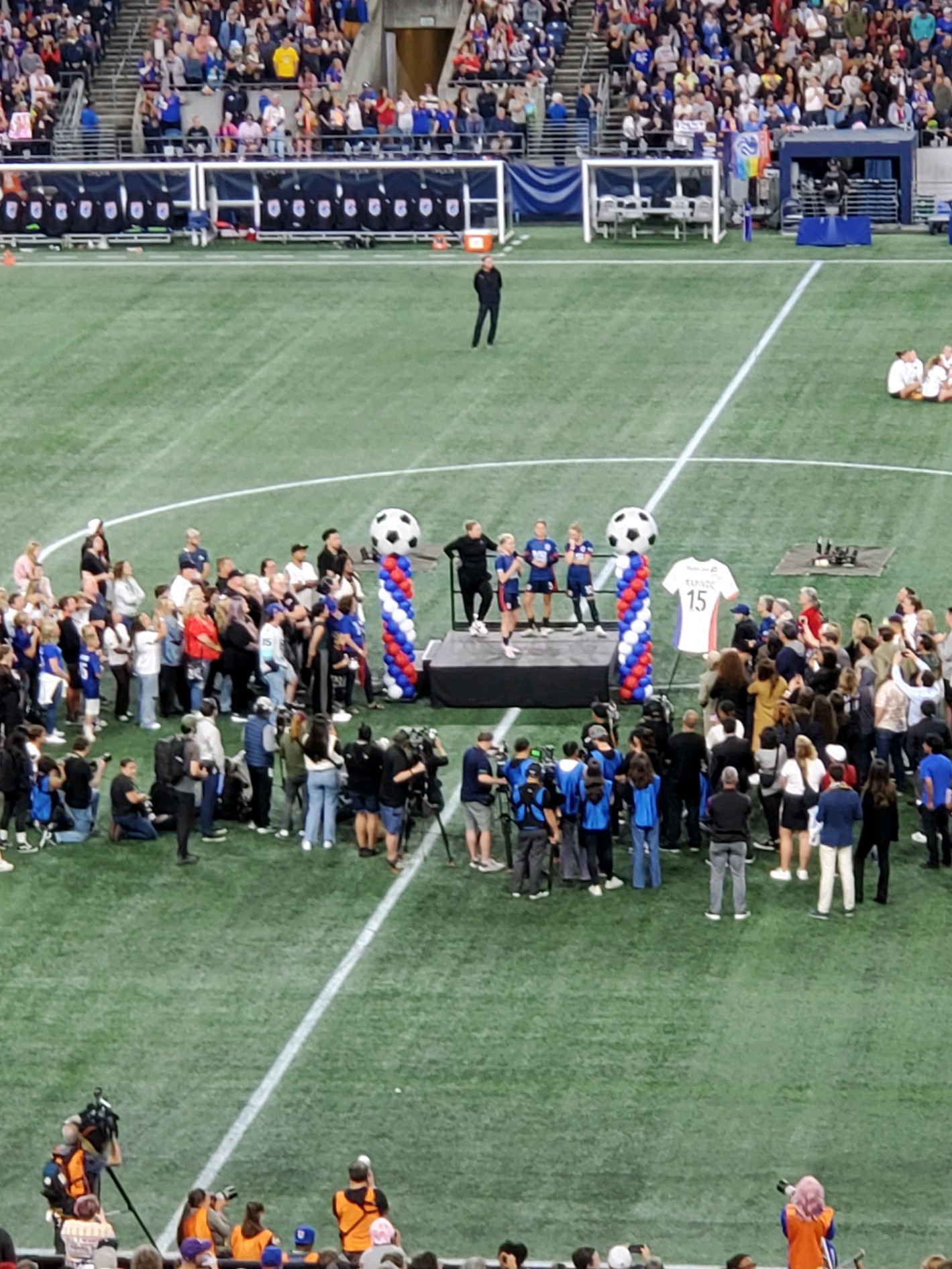 Rapinoe, Fishlock, Barnes, and Harvey standing on a small stage in the middle of a soccer field, surrounded by players, family, and press.