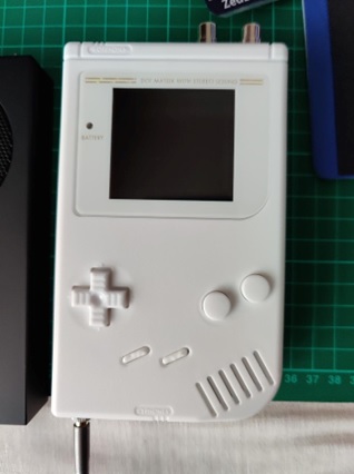 A white Game Boy with two metal sockets added to the top right of the console, and another socket on the bottom left with a lead attached to it.