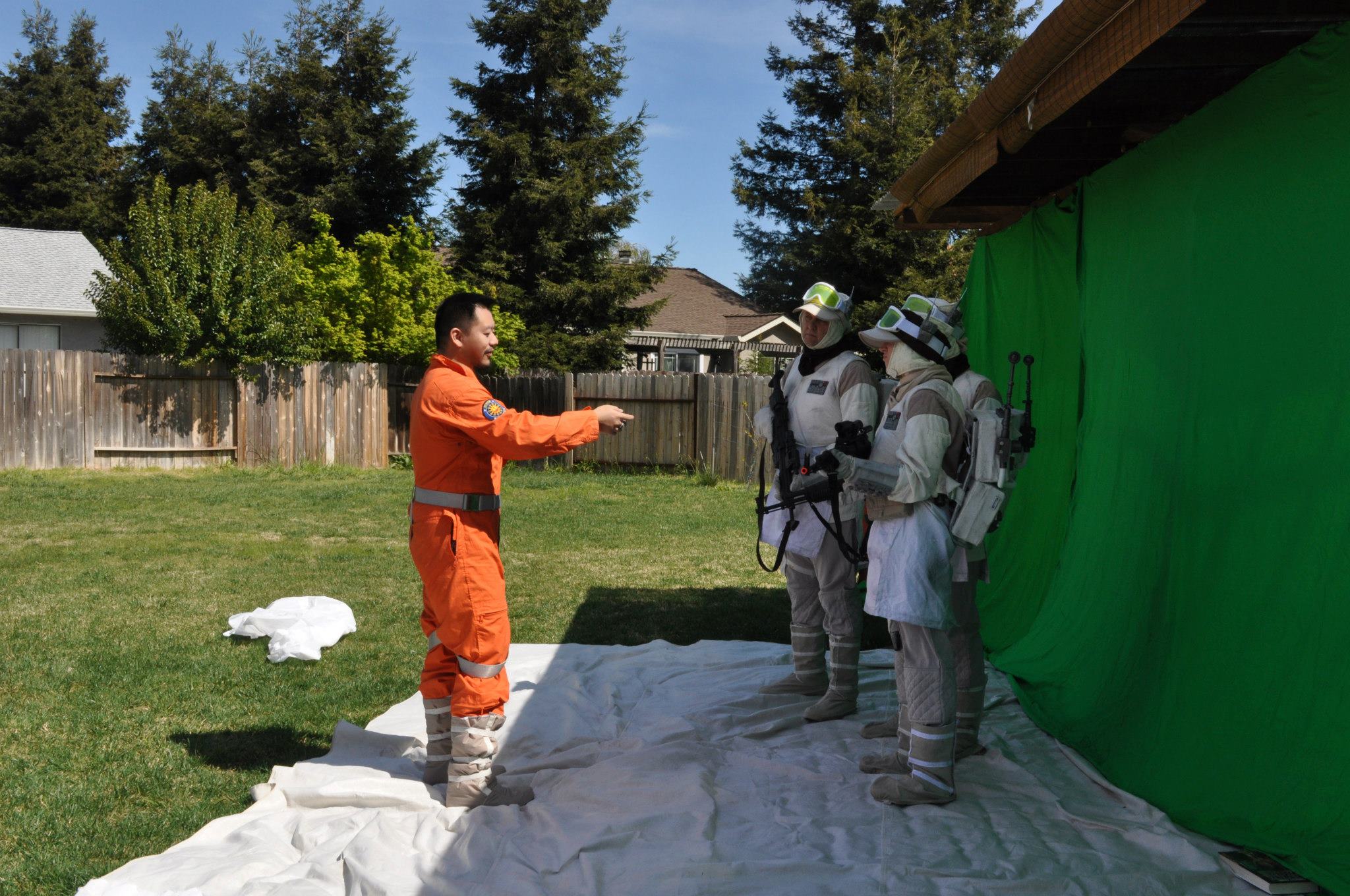 Color photo of man in orange jumpsuit directing two men dressed in rebel soldier uniforms standing in front of a green screen in a backyard.