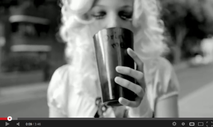 Black-and-white image of an out-of-focus blonde woman, eyes closed, raising an opaque glass/cup reading 'drink me' to her lips. The glass/cup is in focus.